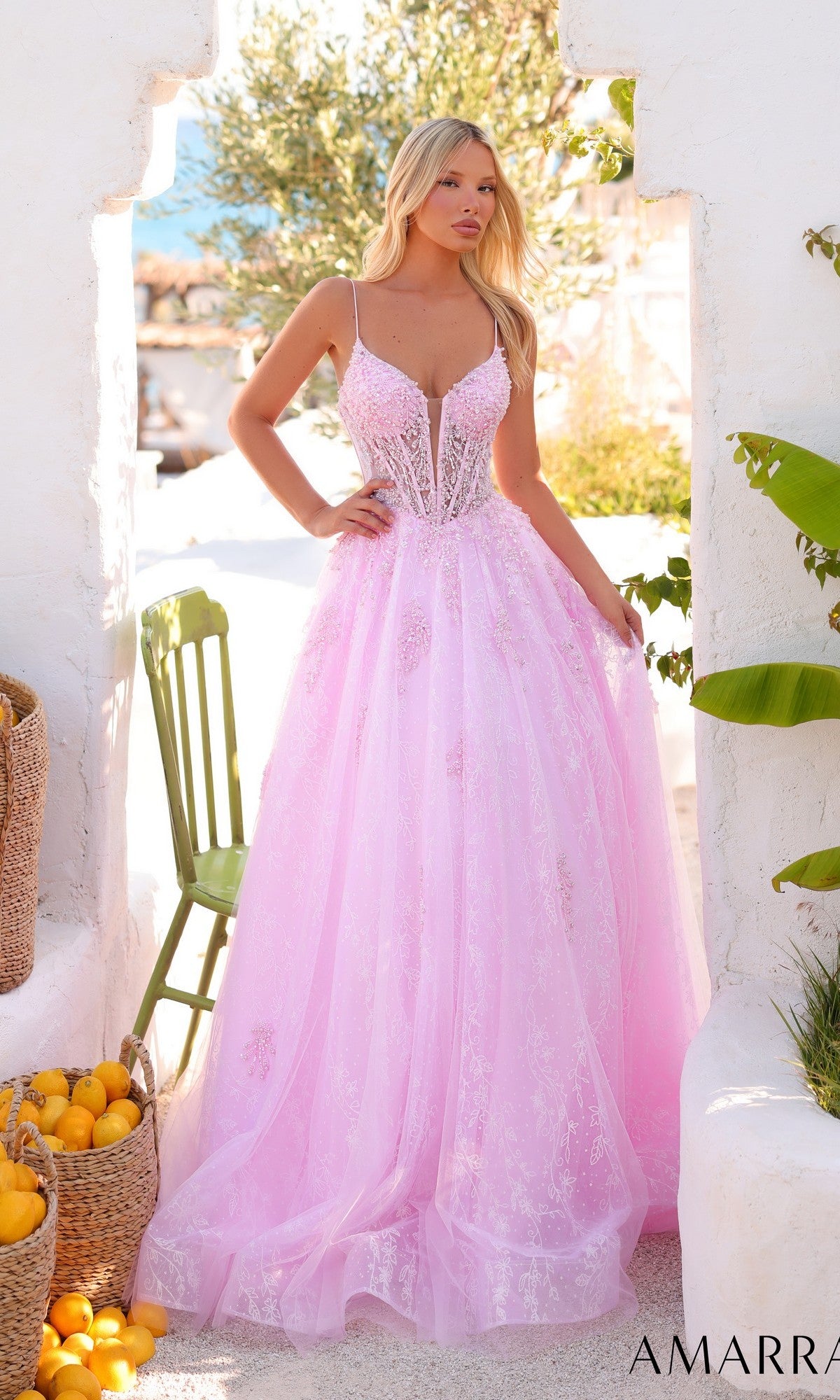 A woman stands outdoors, surrounded by baskets of lemons and white stone walls, wearing the Amarra 89425 long prom dress—a pale pink, floor-length gown with thin straps and a sheer corset bodice.